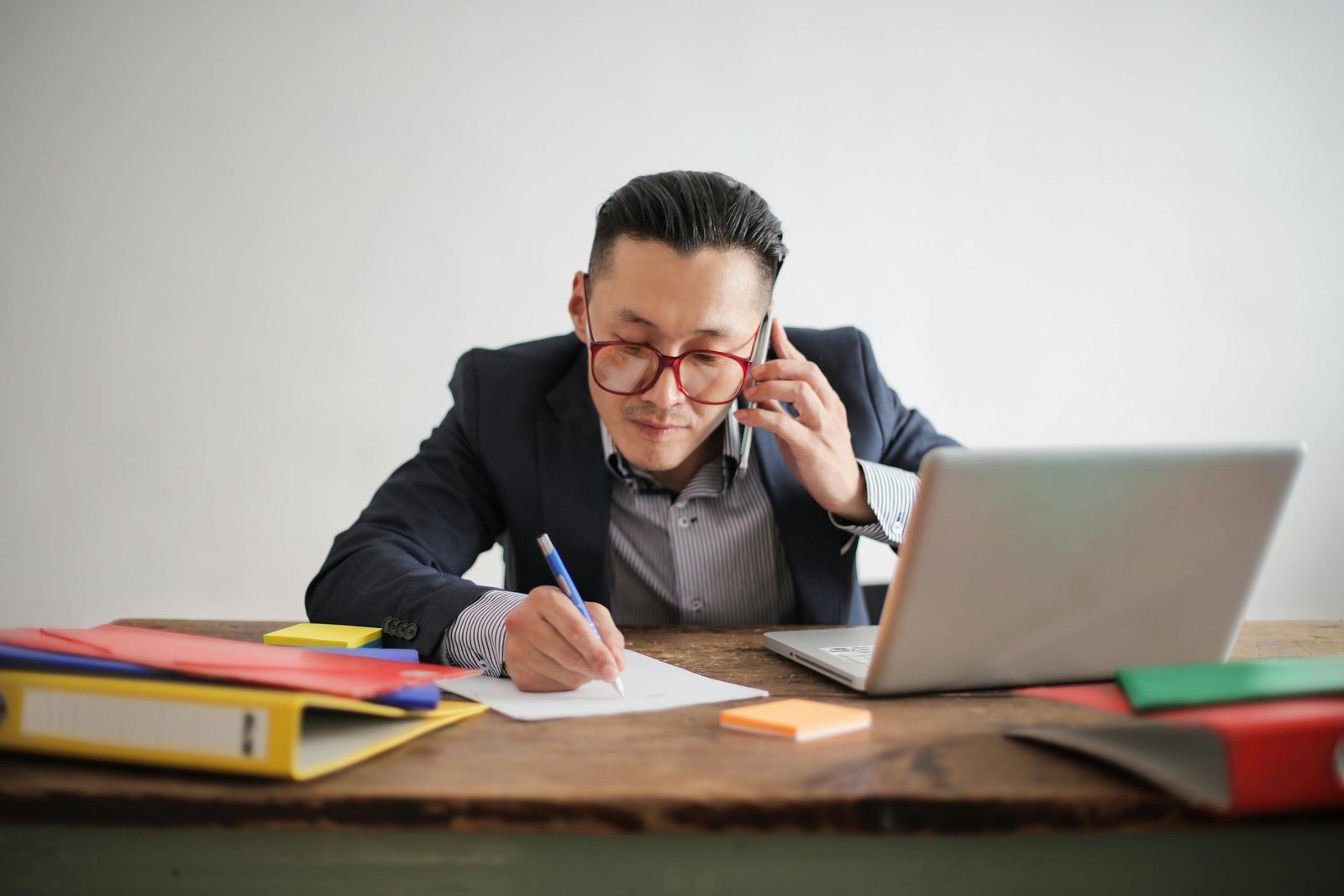 Professional businessman multitasking with phone call and laptop while writing notes at desk.